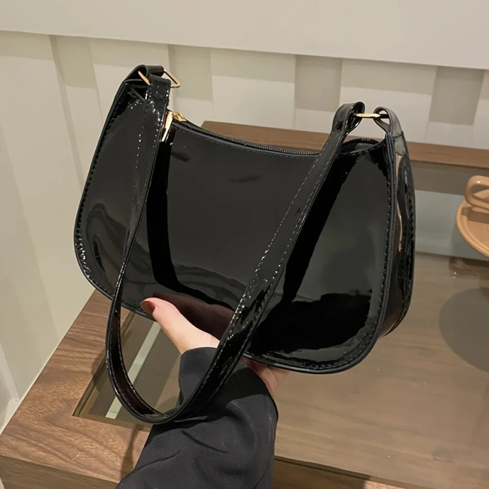 A glossy black patent leather shoulder bag with gold-tone hardware, held by a hand over a wooden table.